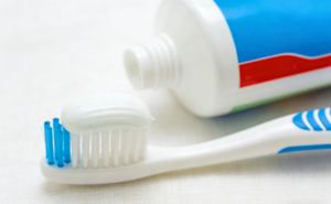 Close-up of white toothpaste with blue specks on a white and blue manual toothbrush, with a toothpaste tube in the background on a white towel